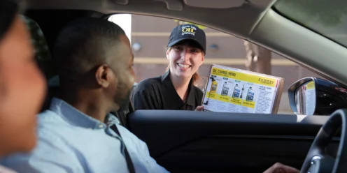 Uncle Ed's Oil Shoppe employee offering oil change services to a customer in a vehicle. The technician holds an oil change menu and provides professional automotive maintenance advice in a friendly manner.