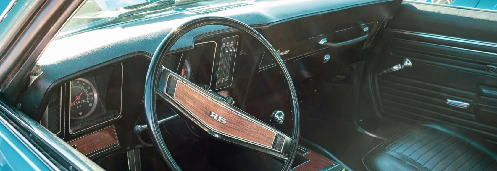 Interior of a classic car from the Cars of Character collection, featuring a black dashboard, wood-trimmed steering wheel with "RS" emblem, manual gear shift, and sleek black leather seats.