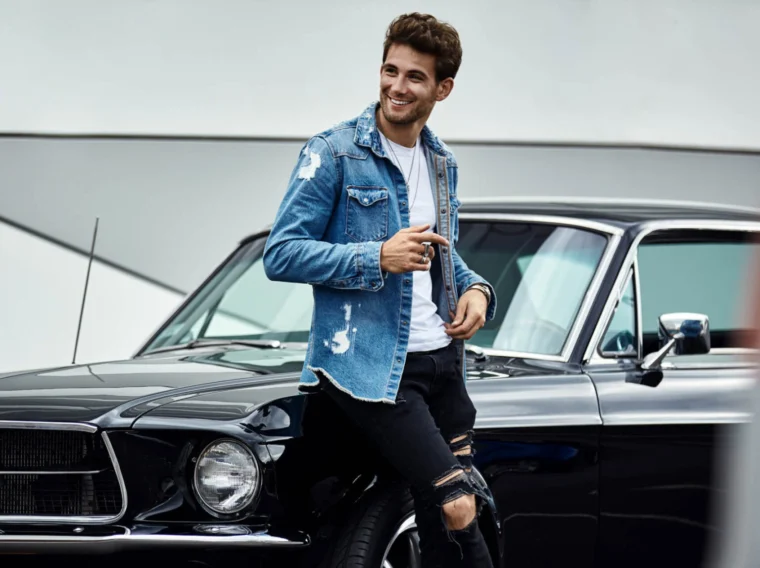 A man in a ripped denim jacket and black jeans leans against a vintage black car from Cars of Character, smiling and holding sunglasses.