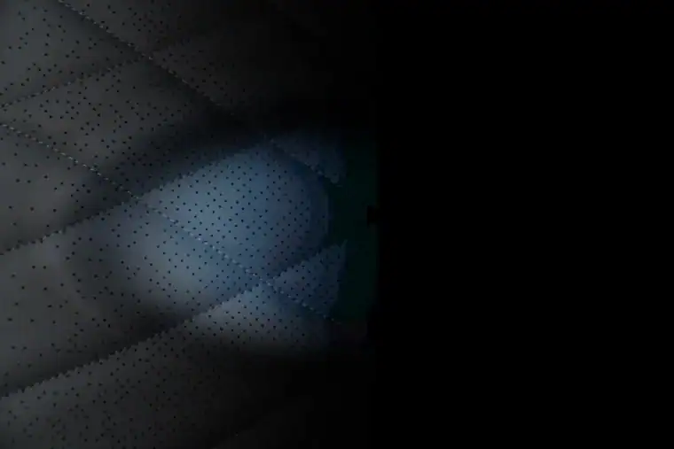 Close-up of a quilted, perforated surface partially illuminated, with the rest fading into darkness on the right side of the image.