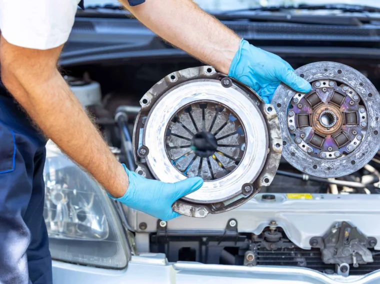 Auto mechanic holding used car pressure plate and clutch disc in front of the vehicle engine.