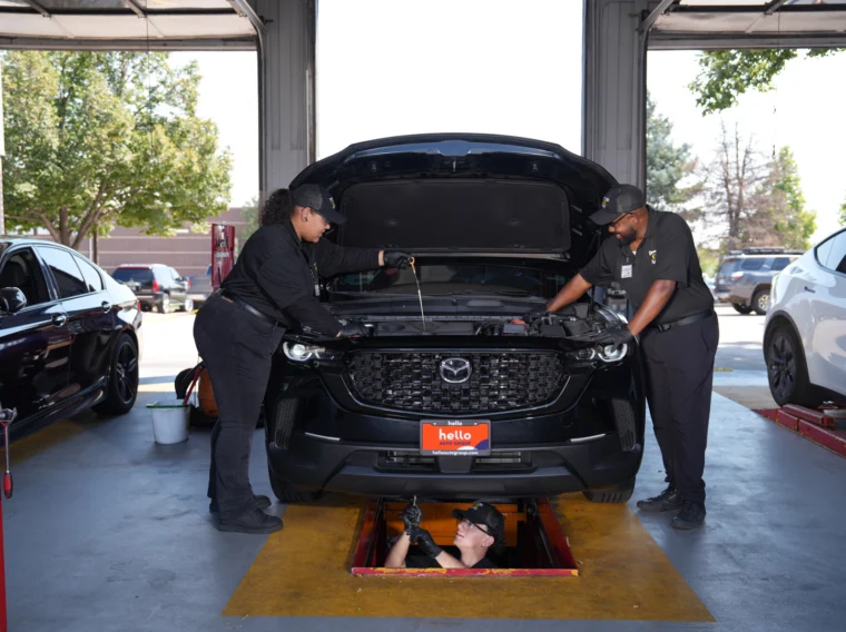 Two mechanics are working on a black Mazda SUV with its hood open, while a third person inspects the vehicle from an underground service bay below.