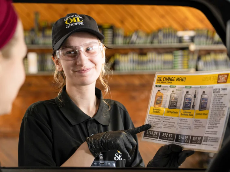 An employee wearing gloves and safety glasses shows a customer an oil change menu through a car window, pointing at the options listed on the laminated sheet.