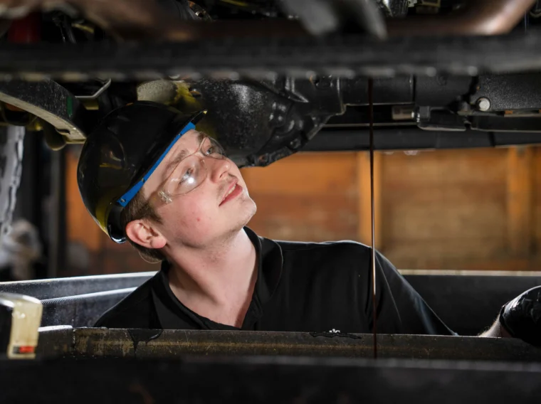 A mechanic wearing safety glasses and a hard hat inspects the underside of a vehicle as oil drains into a container below.