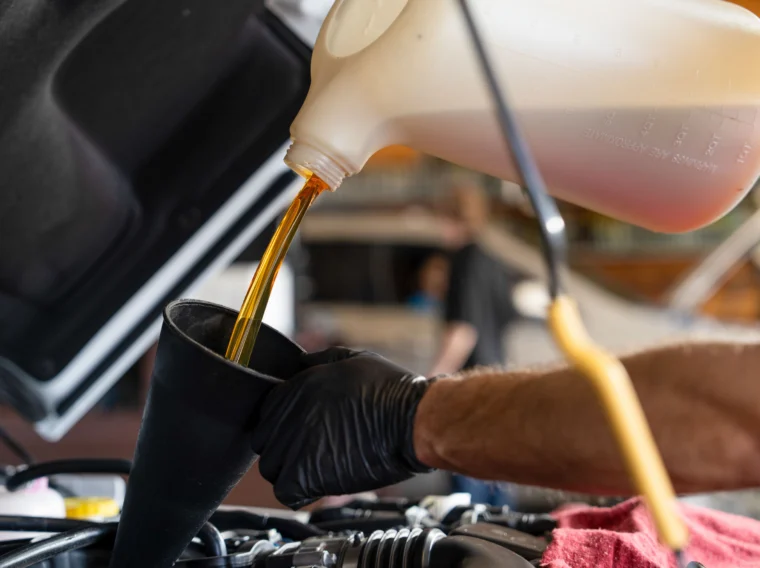 A person wearing black gloves pours motor oil from a plastic container into a funnel under a car hood.