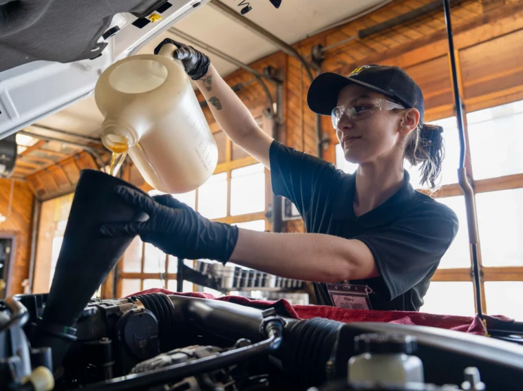A mechanic wearing gloves and safety glasses pours oil into a car engine using a funnel in a well-lit garage.