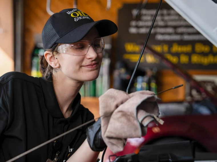 A mechanic wearing safety glasses and a cap is wiping an engine dipstick with a cloth while inspecting a car in a garage.