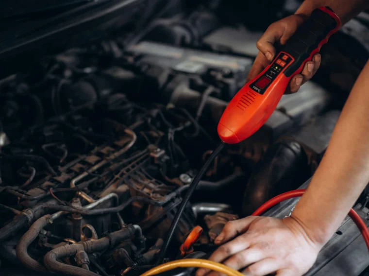 A person uses a red diagnostic tool under the hood of a car, checking wires and engine components while performing AC repair or air conditioning services.