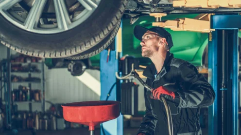 Mechanic in work clothes and gloves stands under a raised car, inspecting the underside and preparing to perform maintenance in an auto repair shop.