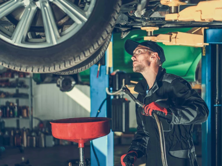 Mechanic in work clothes and gloves stands under a raised car, inspecting the underside and preparing to perform maintenance in an auto repair shop.