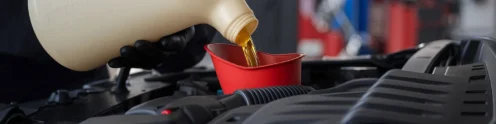 A person wearing black gloves pours engine oil from a large container into a red funnel placed in a car engine bay.