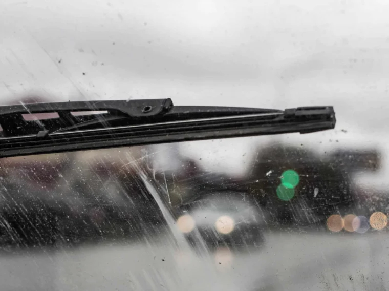 Close-up of a windshield wiper moving across a dirty windshield on a rainy day, with blurred traffic lights and cars in the background—highlighting the importance of regular wiper replacement services for clear visibility.