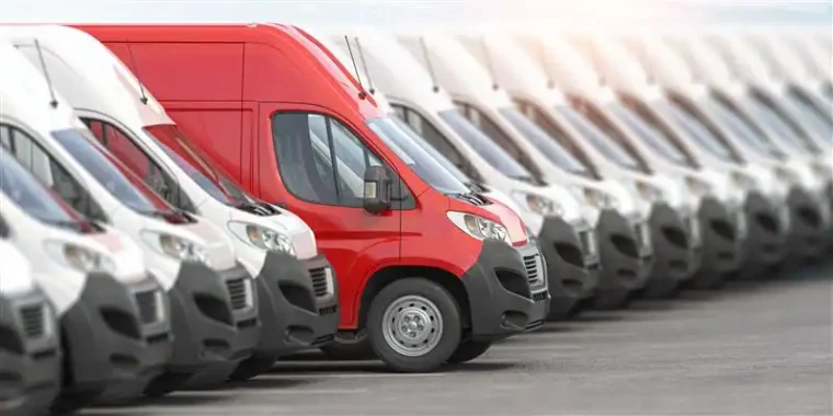 A row of white vans is parked in a lot, with one red van standing out among them.