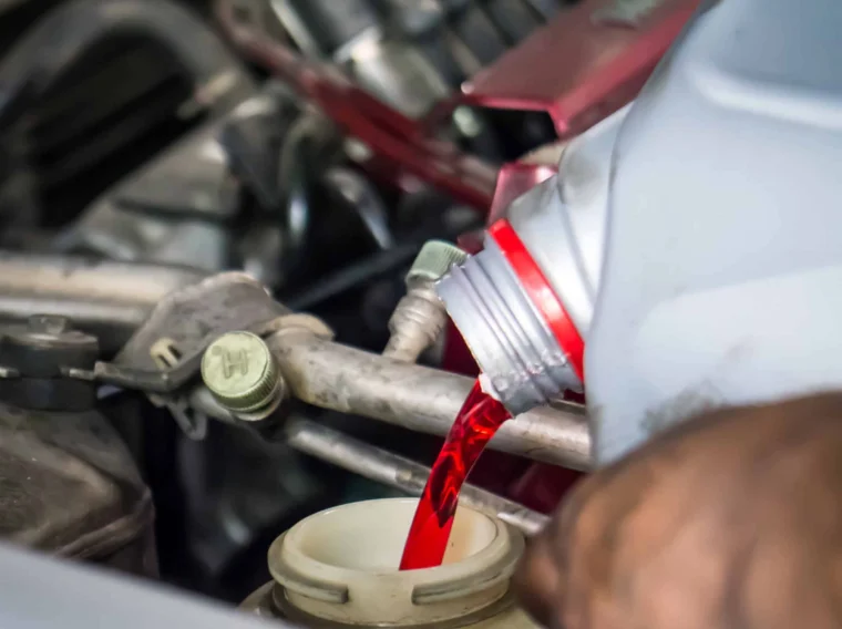 A person pours red fluid from a plastic bottle into a car engine compartment, likely adding transmission or power steering fluid.