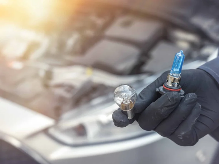 A gloved hand holds two car headlight bulbs, one traditional and one halogen, in front of an open car hood—perfect for showcasing professional Headlight Services.