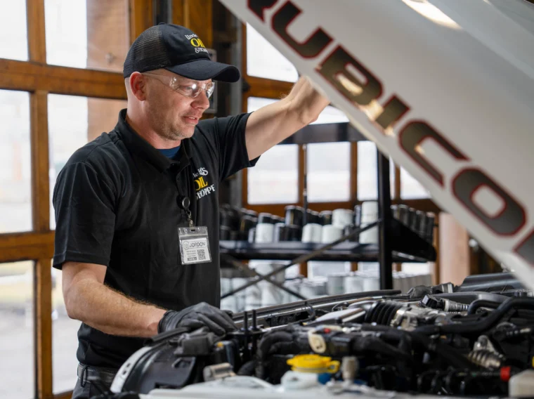 A mechanic wearing a cap and name badge inspects the engine of a vehicle with the hood open inside a garage, checking for maintenance needs such as Transmission Fluid Services.