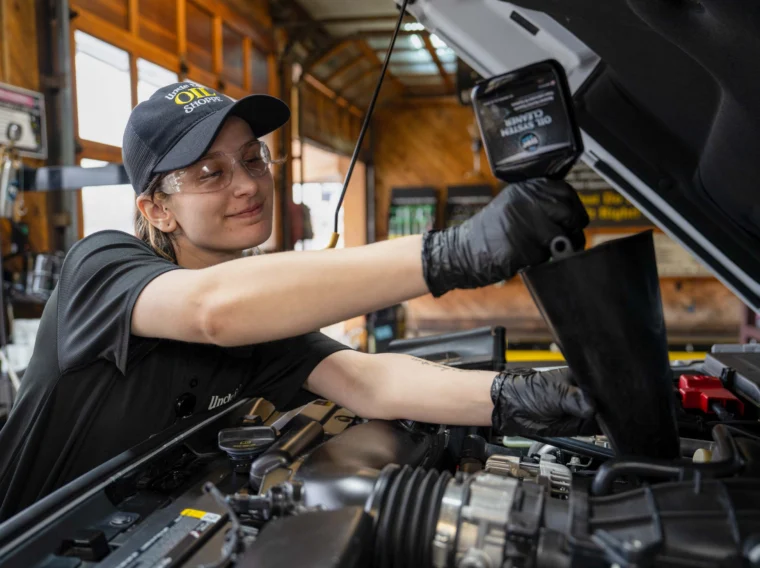 A mechanic wearing gloves and safety glasses pours motor oil into a car engine, performing essential engine maintenance in an auto repair shop.