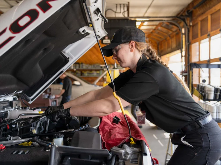 A mechanic wearing gloves and safety glasses works under the hood of a vehicle, performing power steering services inside an auto repair shop.