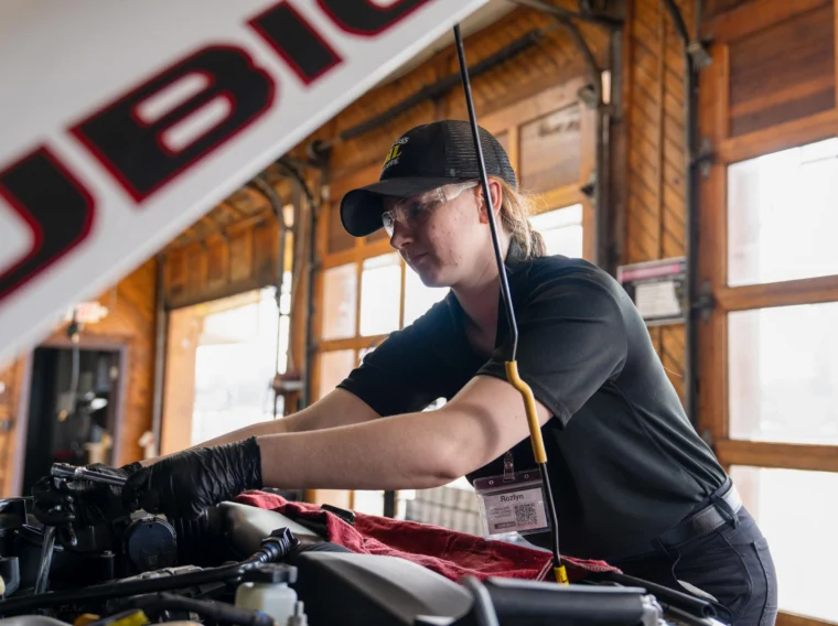 A mechanic wearing gloves and a cap performs engine maintenance under the hood of a vehicle inside a garage with large windows.