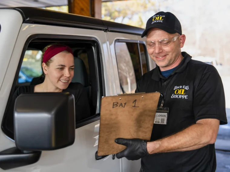 Oil shop employee in uniform shows a clipboard to a woman sitting in her car, both smiling, during an engine treatment service interaction.