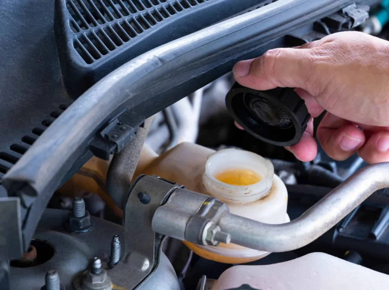 A person’s hand unscrews the cap of a brake fluid reservoir under a car hood, exposing the yellowish fluid inside.