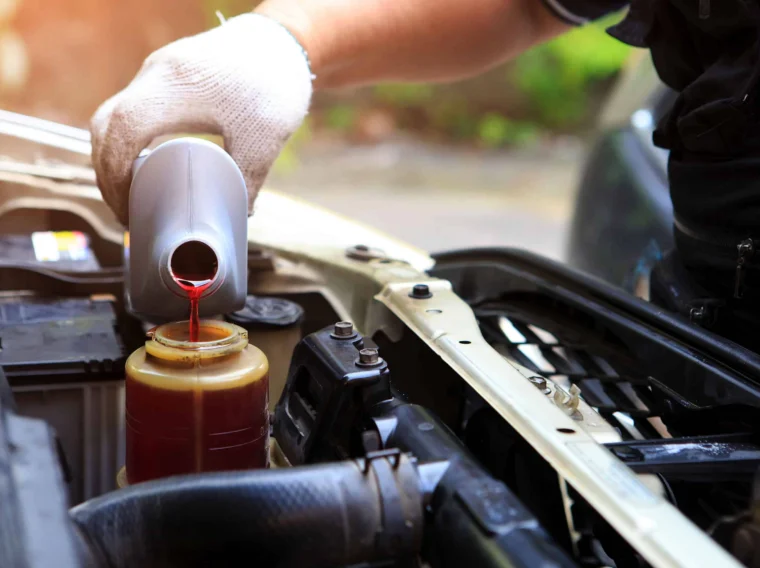 A person wearing a white glove pours red automotive fluid from a gray bottle into a reservoir under a car hood, demonstrating essential steering maintenance.