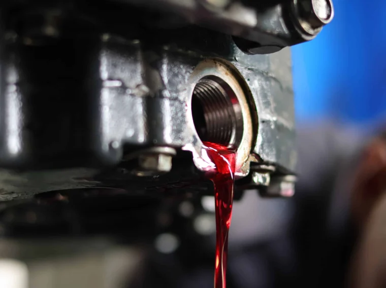 Red fluid drains from the threaded opening of a metal engine component, suggesting an oil or transmission fluid change in progress.