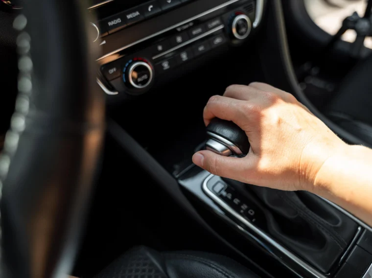 A person’s hand is gripping the gear shift of an automatic car, with the dashboard and controls visible in the background.