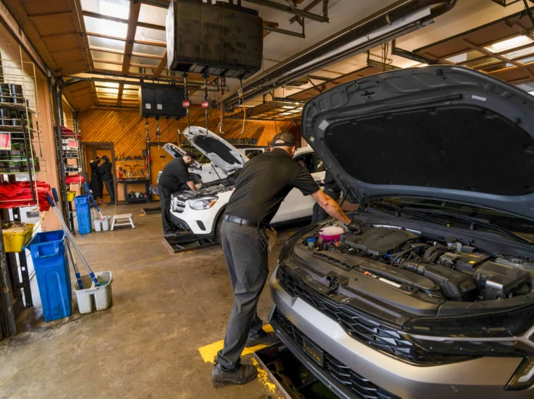 Two mechanics provide installation services as they work on cars with open hoods inside a garage, using tools and equipment, with cleaning supplies visible on the left.