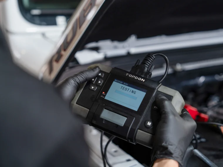 A person wearing black gloves uses a handheld diagnostic device to test a car battery under the open hood of a vehicle, highlighting the importance of routine checks as part of comprehensive cleaning services.