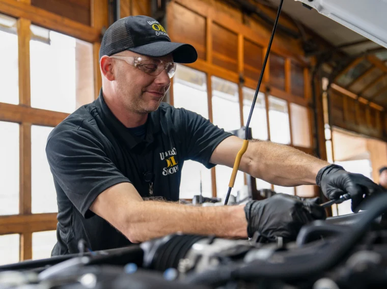 A mechanic wearing safety glasses and gloves works under the hood of a car, providing installation services inside a garage with wooden doors.
