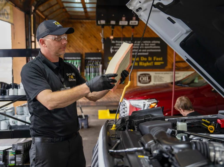A mechanic wearing gloves and a cap inspects an air filter beside an open car hood in an auto service garage, highlighting their expert installation services.