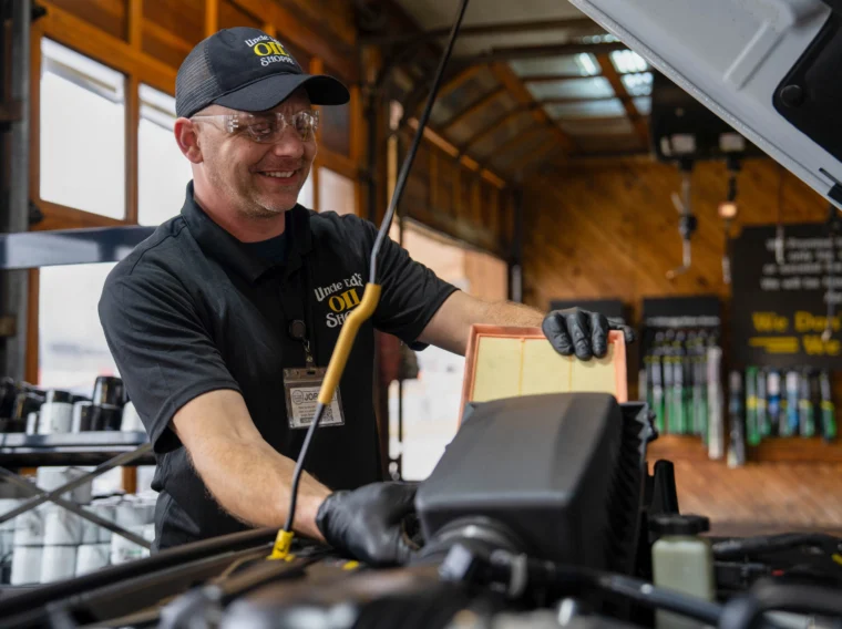 A mechanic wearing gloves and safety glasses checks an air filter under a car hood, providing expert installation services at an auto service shop.