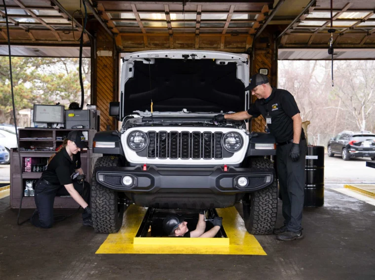 Three mechanics perform Preventive Maintenance Services on a Jeep in the garage—one inspects the engine bay, another works beneath the vehicle, while a third focuses near the front wheel.