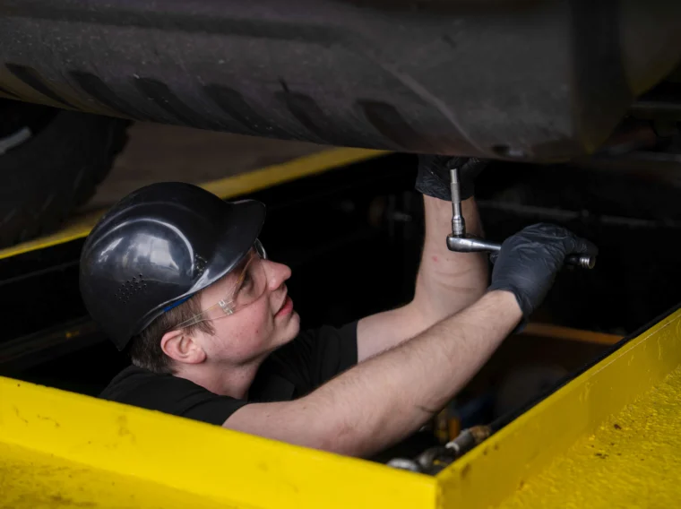 A mechanic wearing safety glasses, gloves, and a hard hat works with a ratchet on the underside of a vehicle while standing in a yellow service pit, ensuring safety and cleanliness similar to professional cleaning services.