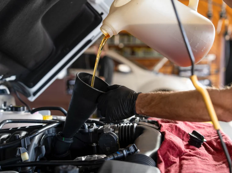 A person wearing black gloves pours motor oil from a large container into a funnel placed in a car engine bay, ensuring the area stays neat—just like you’d expect from top-tier cleaning services.