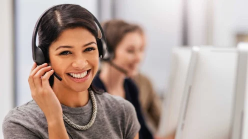 A woman wearing a headset smiles at the camera while sitting at a desk in an office, with another person working at a computer in the background.