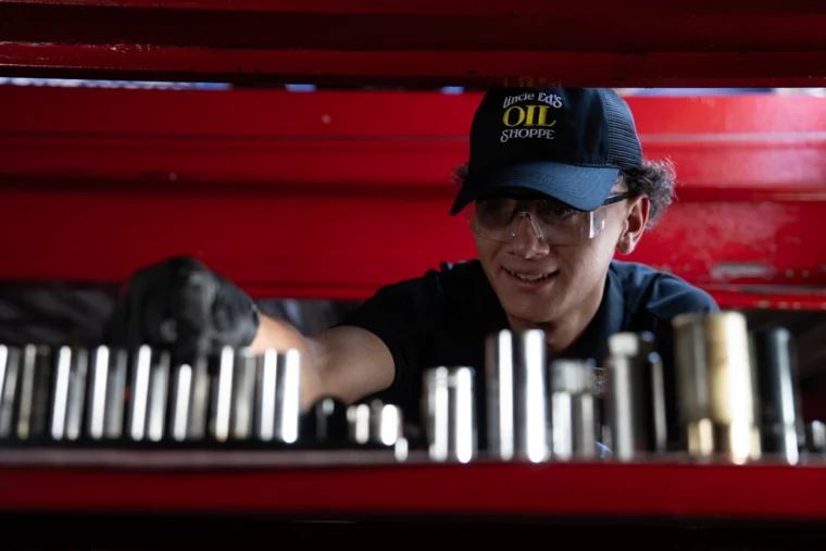 A person wearing safety glasses and a "Uncle Ed's Oil Shoppe" hat selects a socket from a row of metal tools in a workshop with red shelving.