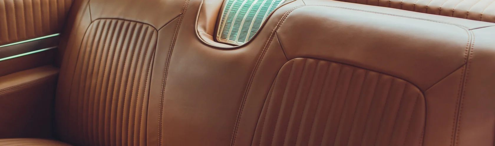 Close-up of a Cars of Character classic car’s brown leather backseat with vertical stitching and a metal air vent centered on the rear deck.