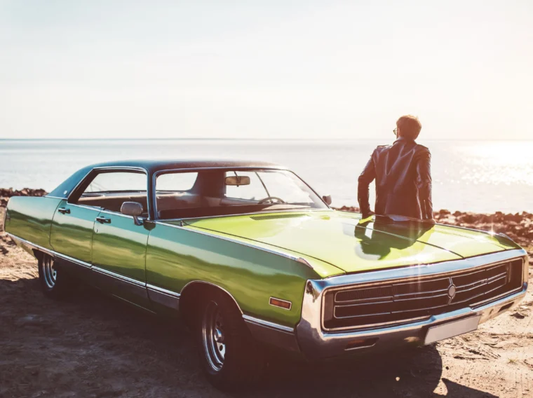 A person stands beside a vintage green car—one of those true Cars of Character—parked on a beach, gazing at the water as sunlight glimmers in the background.