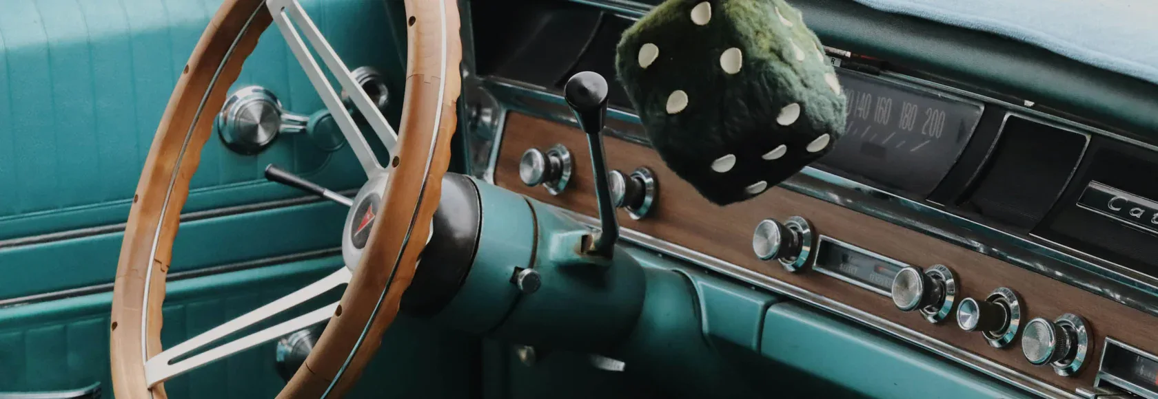 Interior of a vintage teal car from Cars of Character, featuring a wooden steering wheel, manual gear shift, retro dashboard controls, and fuzzy dice hanging from the rearview mirror.