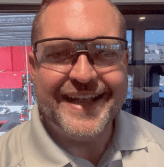 A man wearing safety glasses and a light gray collared shirt smiles at the camera indoors, with a workshop visible in the background.