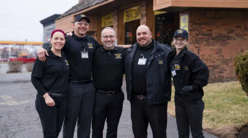 Five people in matching work uniforms stand together and smile outside a brick building on an overcast day.