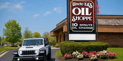 A white Jeep is parked in front of Uncle Ed’s Oil Shoppe, which advertises a 10-minute oil change on its sign; flowers and bushes line the driveway.