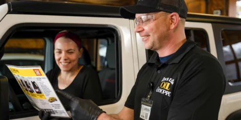 A mechanic wearing gloves and safety glasses shows an oil change brochure to a woman sitting in a vehicle.