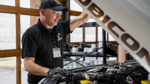 A mechanic wearing a cap and gloves inspects the engine of a white Jeep Rubicon inside an auto repair shop.