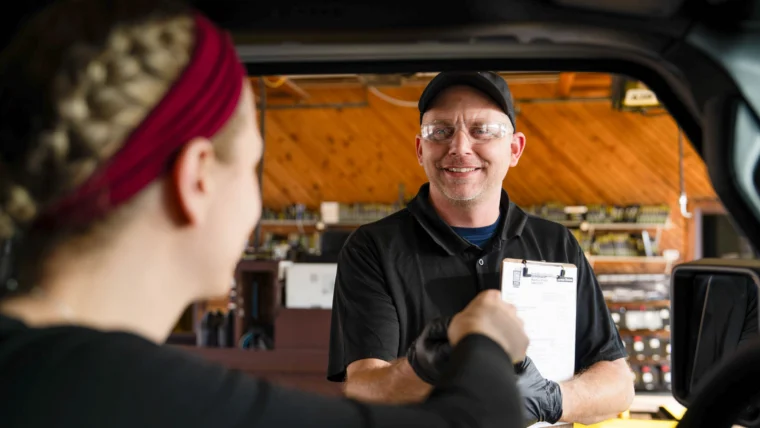 A mechanic holding a clipboard shakes hands with a customer sitting in a vehicle inside an auto repair shop.
