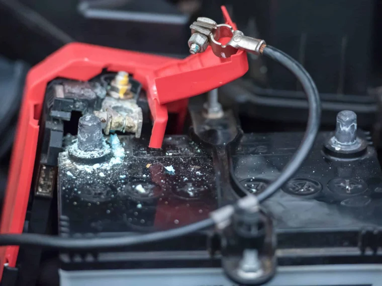 Close-up of a car battery with corrosion and white residue around the positive terminal; a red protective cover is partially open, highlighting the need for professional Battery Services.