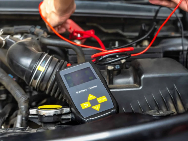 A person uses a battery tester with yellow buttons to check a car battery under the hood, connecting the device with red and black cables as part of routine Battery Services.