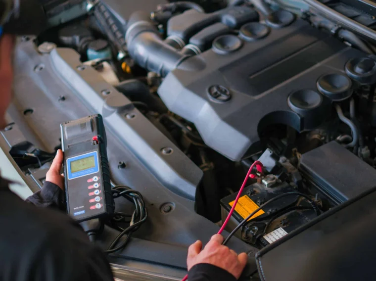 A person uses a handheld diagnostic tool to check a car battery under the open hood of a vehicle in a garage, demonstrating professional Battery Services.